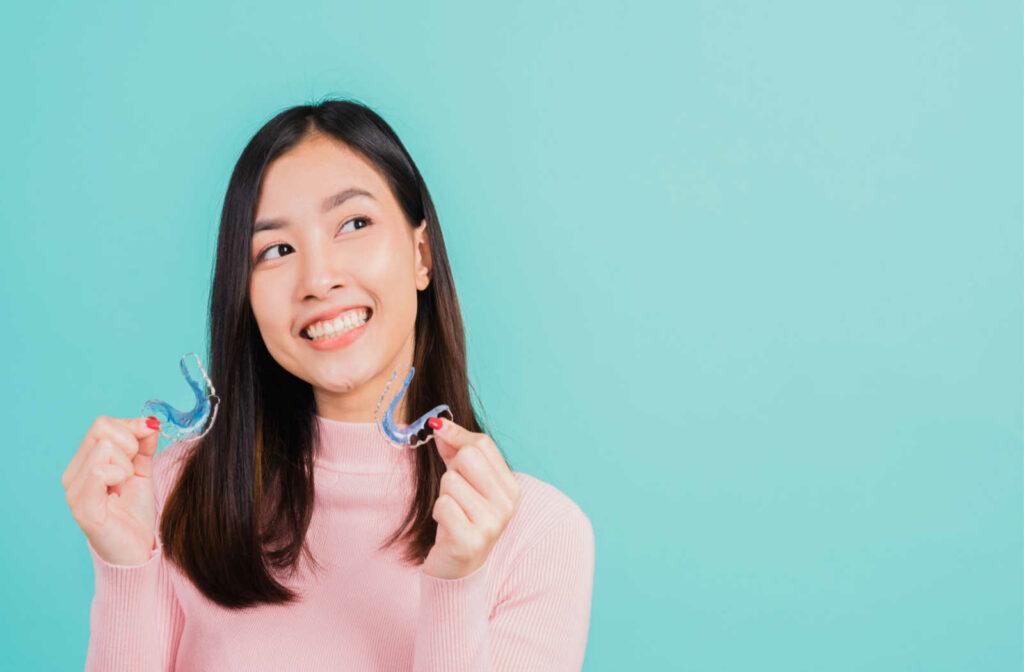 A smiling woman standing against a turquoise background holds two retainers.