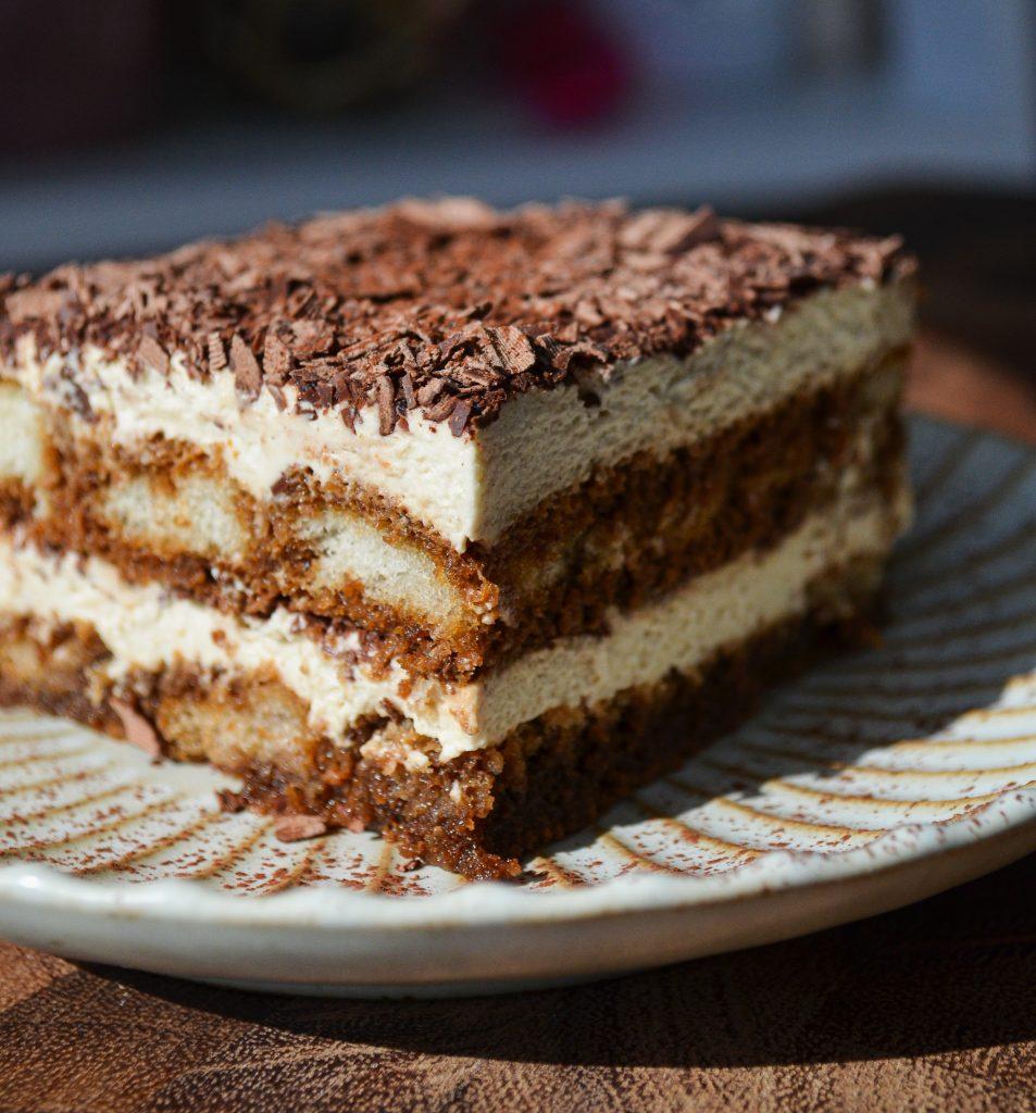 tiramisu with layers of chocolate shavings, mascarpone cheese and soaked ladyfingers sitting on a plate with dusted powder